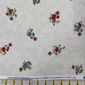 A close-up of patterned fabric with a white base, fine tan lattice grid, and scattered floral motifs in red, blue, green, and small red-and-blue striped accents. A ruler strip is visible along the bottom edge.